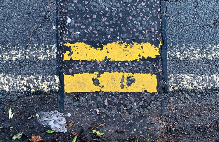 A photograph of a tarmac street with two yellow lines. Where a repair has been made to the road surface, the yellow lines are a brighter yellow. In the bottom of the image, is what looked like a small piece of wet white paper, and green and brown leaves.